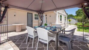 Un patio al aire libre con una mesa, sillas, una sombrilla y una casa al fondo.