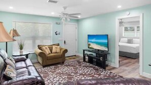 Una sala de estar con un sofá de cuero, un sillón color beige, un televisor sobre un mueble bajo, un ventilador de techo y un tapete estampado.