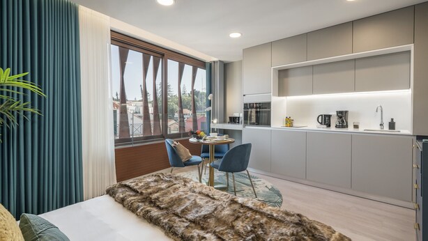 Habitación de hotel moderna con una cama grande, vistas de la ciudad, una pequeña cocina y una mesa de comedor con sillas.