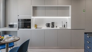 Una cocina moderna con electrodomésticos integrados, una zona de comedor con sillas azules y una pequeña mesa con frutas y tazas.