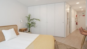 Ein modernes Hotelzimmer mit einem großen Bett, einem Schrank, einem kleinen Tisch mit Pflanze und einem Sessel.