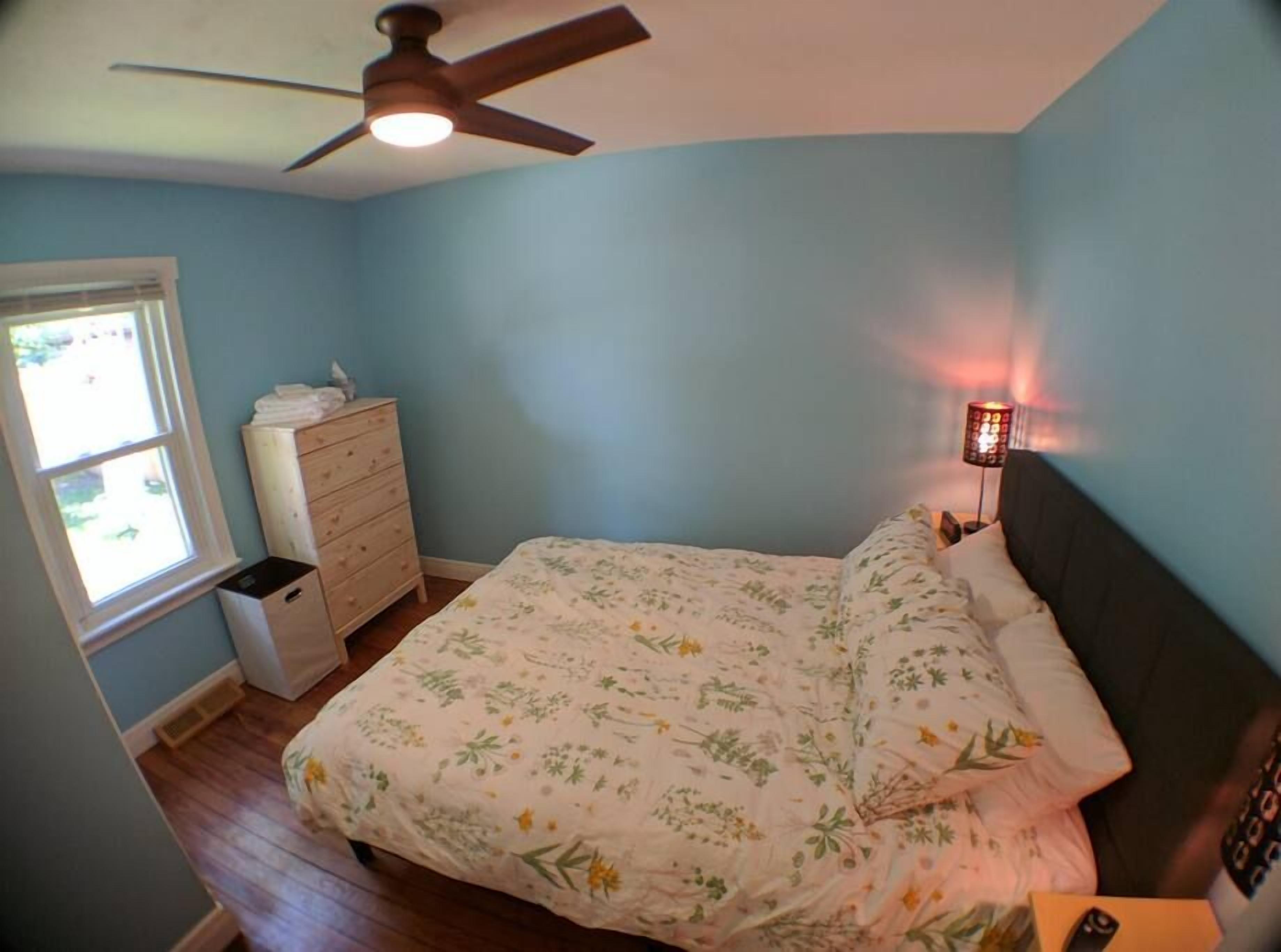 Une chambre à coucher comprenant un lit, une commode, un ventilateur de plafond, une fenêtre et une table de chevet avec une lampe.