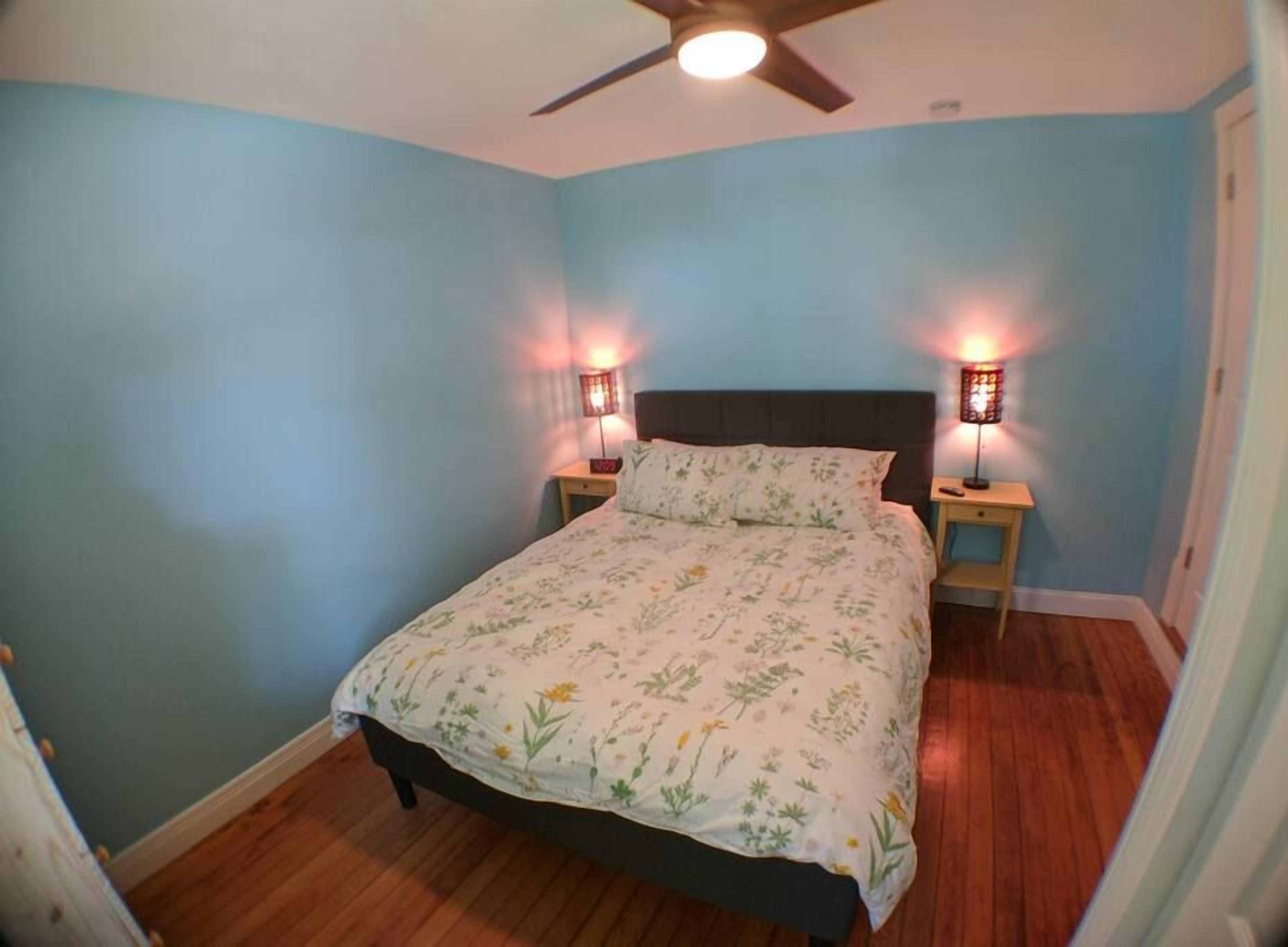 Un dormitorio con cama, dos lámparas de noche, ventilador de techo y suelo de madera.