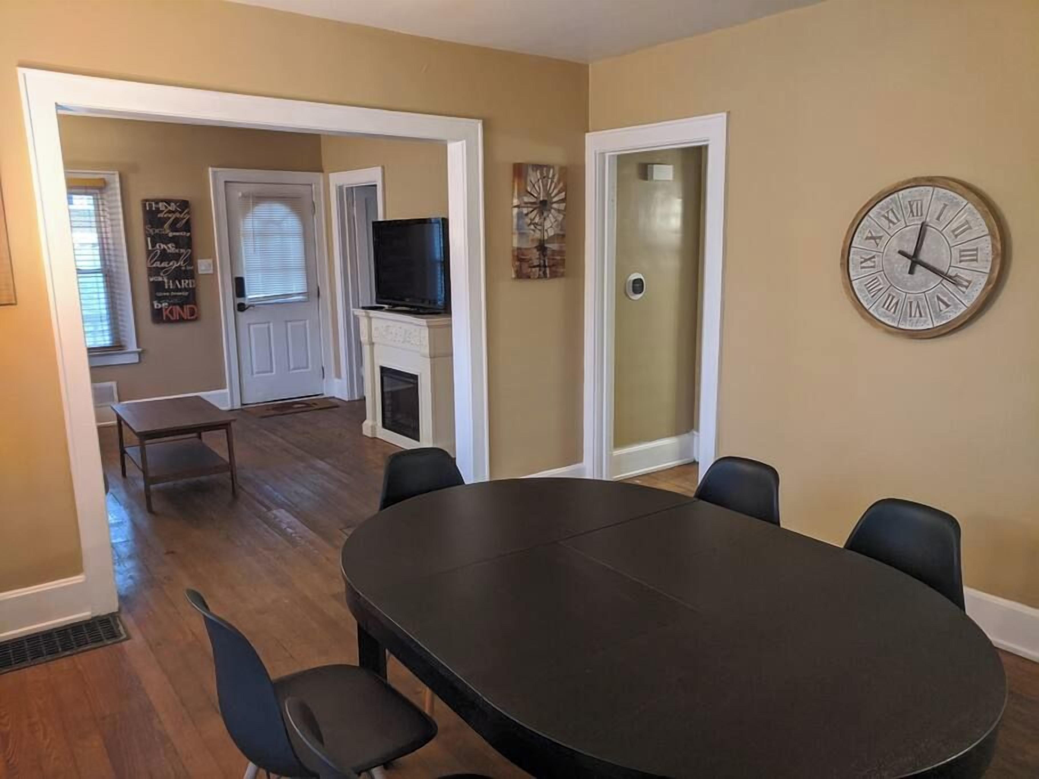 A dining area with a round table, four chairs, a clock on the wall, and a fireplace.