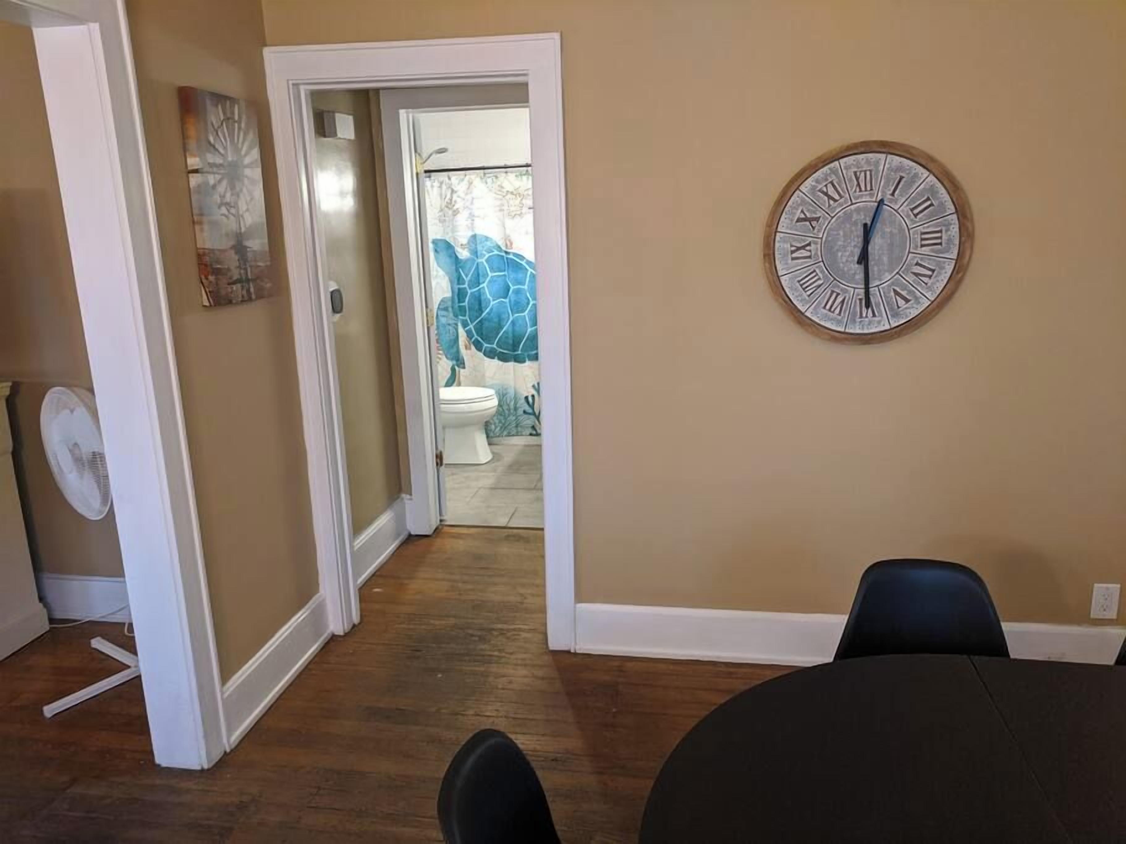 Ein Zimmer mit Holzboden, einer runden Wanduhr, einem Deckenventilator und einem Badezimmer mit Wandmalerei eines blauen Schildkrötenmotivs.