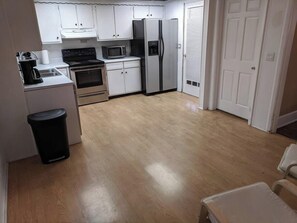 A compact kitchen with white cabinets, a stove, microwave, and refrigerator. A trash bin is visible, and the floor is wooden. - Ohio Ave, 3 bed house, Historic Downtown Loveland (Loveland)