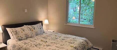 A bedroom with a bed, a nightstand, a lamp, a ceiling fan, and a window with a view of greenery.