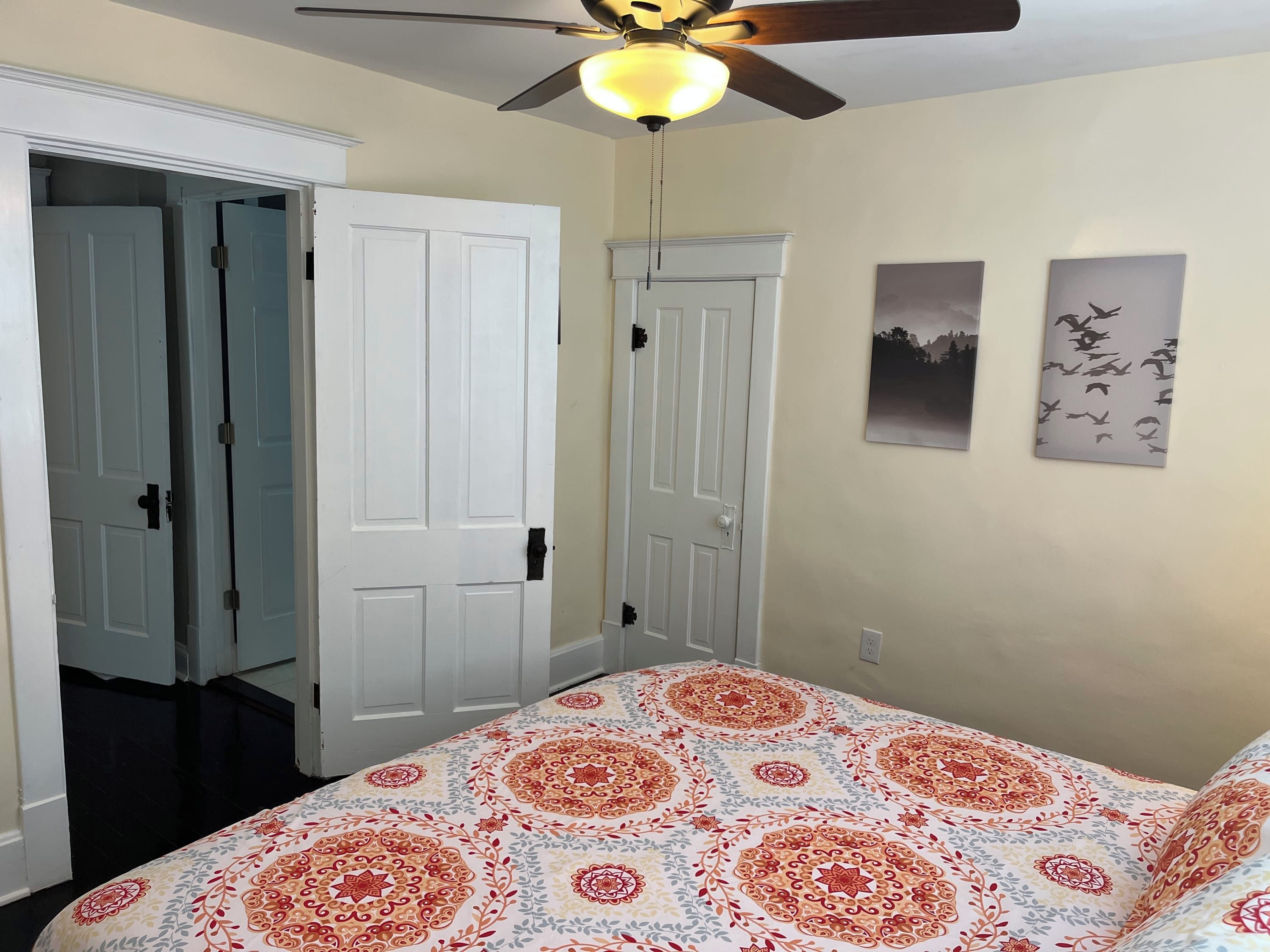 A bedroom with a ceiling fan, two white doors, a patterned bedspread, and two wall art pieces depicting birds.