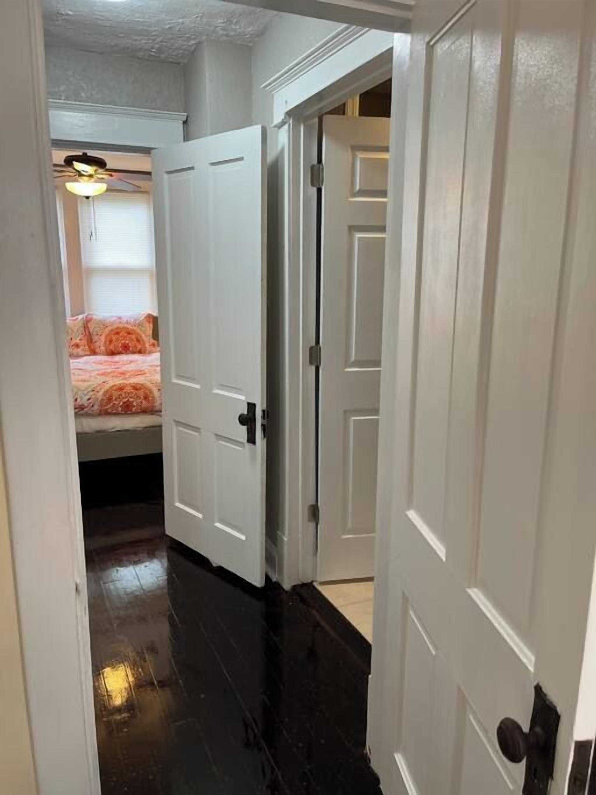 Un couloir étroit, avec des portes blanches donnant sur une chambre à coucher équipée d’un lit et d’un ventilateur de plafond.