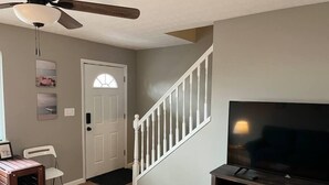A living room with a wooden floor, a white door, a staircase with white railings, a ceiling fan, and a flat-screen TV on a wooden stand. - Carter #4, Two bedroom unit near Xavier, Downtown (Cincinnati)