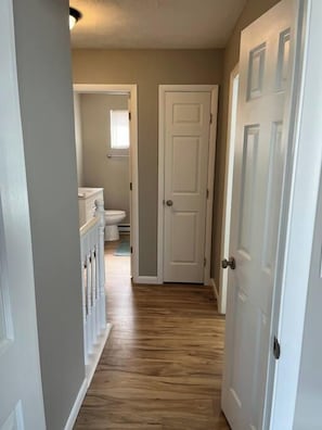 A narrow hallway with wooden flooring leading to a bathroom with a toilet and a window.