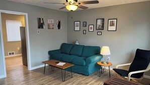 A living room with a teal sofa, a wooden coffee table, a side table with a lamp, a black chair, and a ceiling fan.