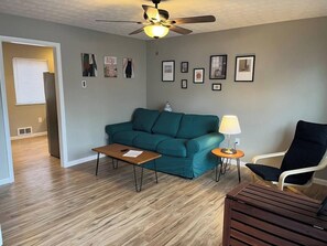 A living room with a teal sofa, a wooden coffee table, a side table with a lamp, a black chair, and a ceiling fan.