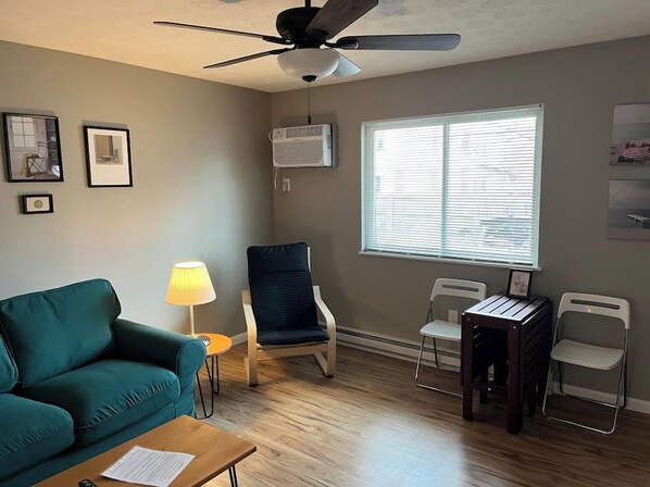 A living room with a teal sofa, a ceiling fan, a window with blinds, and a small table with a lamp.
