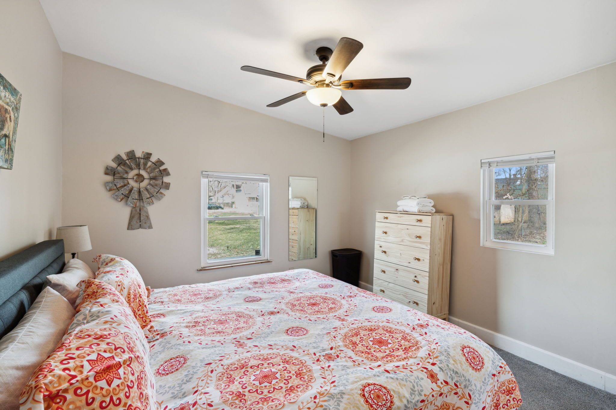 A bedroom with a bed, a ceiling fan, a dresser, a mirror, and two windows.