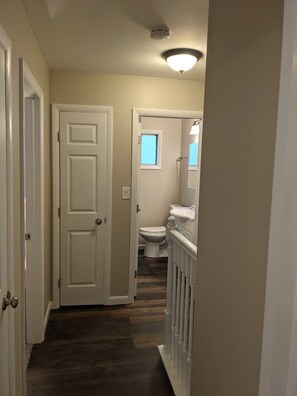 Un couloir étroit avec un plancher en bois, une porte blanche et une salle de bain visible au fond.