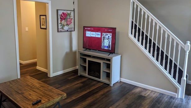A living room with a wooden floor, a TV on a stand, a staircase, and a door leading to another room.