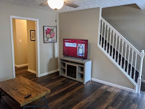 A living room with a wooden floor, a TV on a stand, a staircase, and a door leading to another room. - Carter #5, Private Two Bed near Xavier, Downtown (Cincinnati)