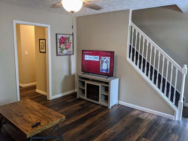 Un salon avec un plancher en bois, un téléviseur posé sur un meuble, un escalier et une porte donnant sur une autre pièce.