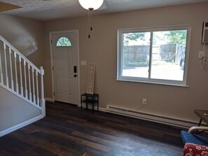 Quarto com piso de madeira, porta branca, janela com vista para um quintal e uma pequena mesa com uma cadeira.