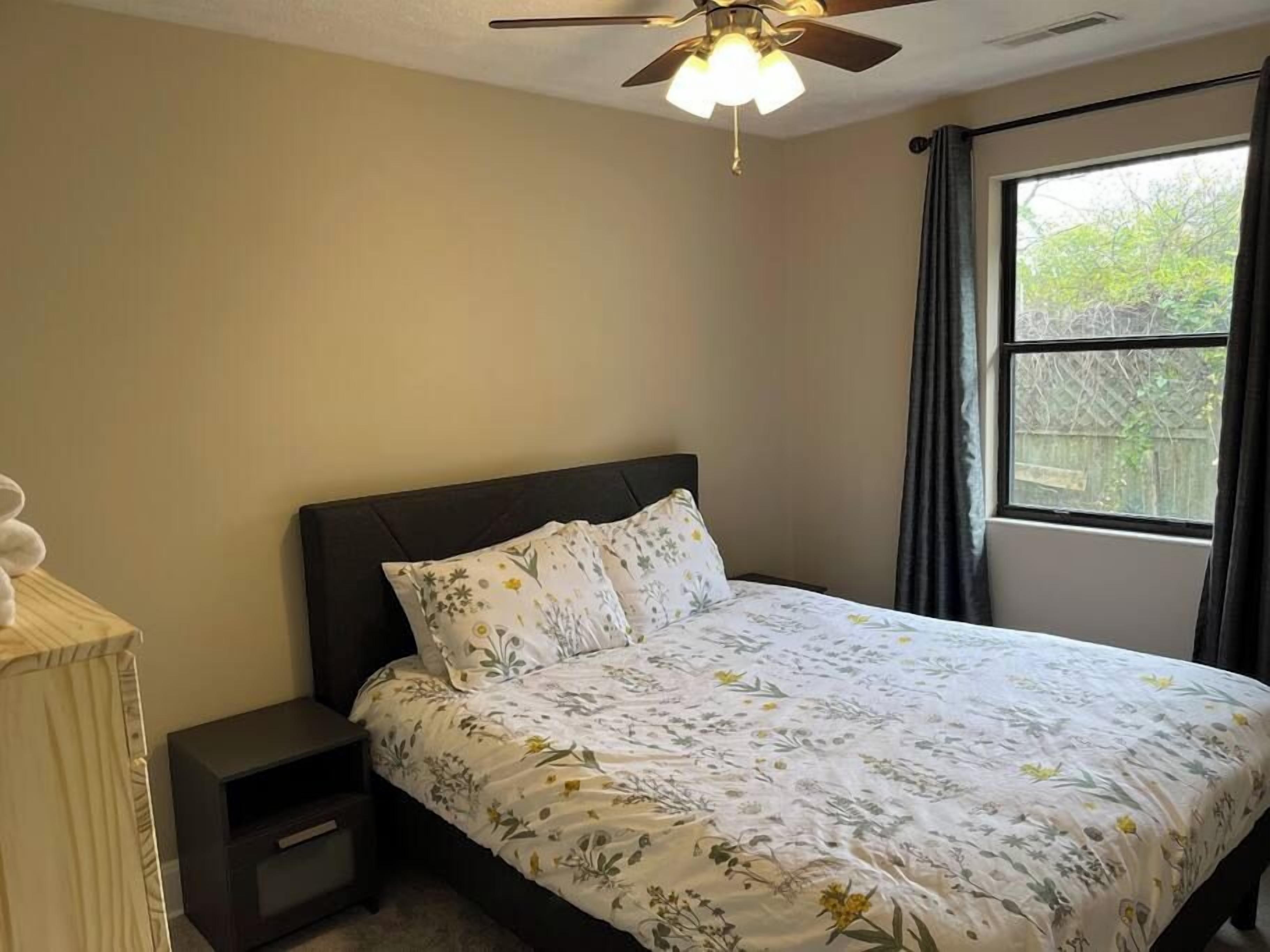 A bedroom with a bed, a nightstand, a ceiling fan, and a window with curtains.