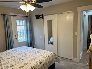 A bedroom with a bed, a ceiling fan, a window with curtains, a door with a mirror, and a hallway leading to another room.
