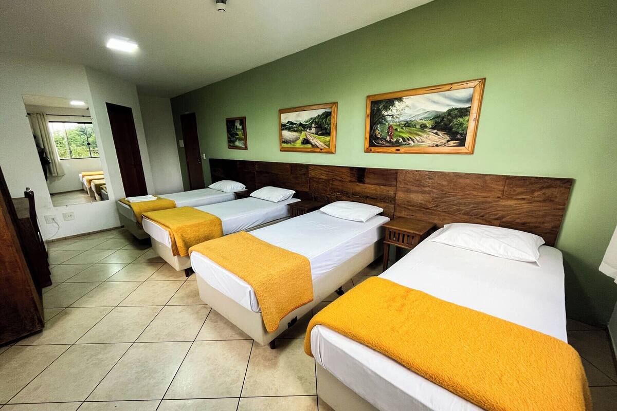 A hotel room with four single beds, each with a white mattress and a yellow blanket. There are two framed landscape paintings on the wall above the beds.