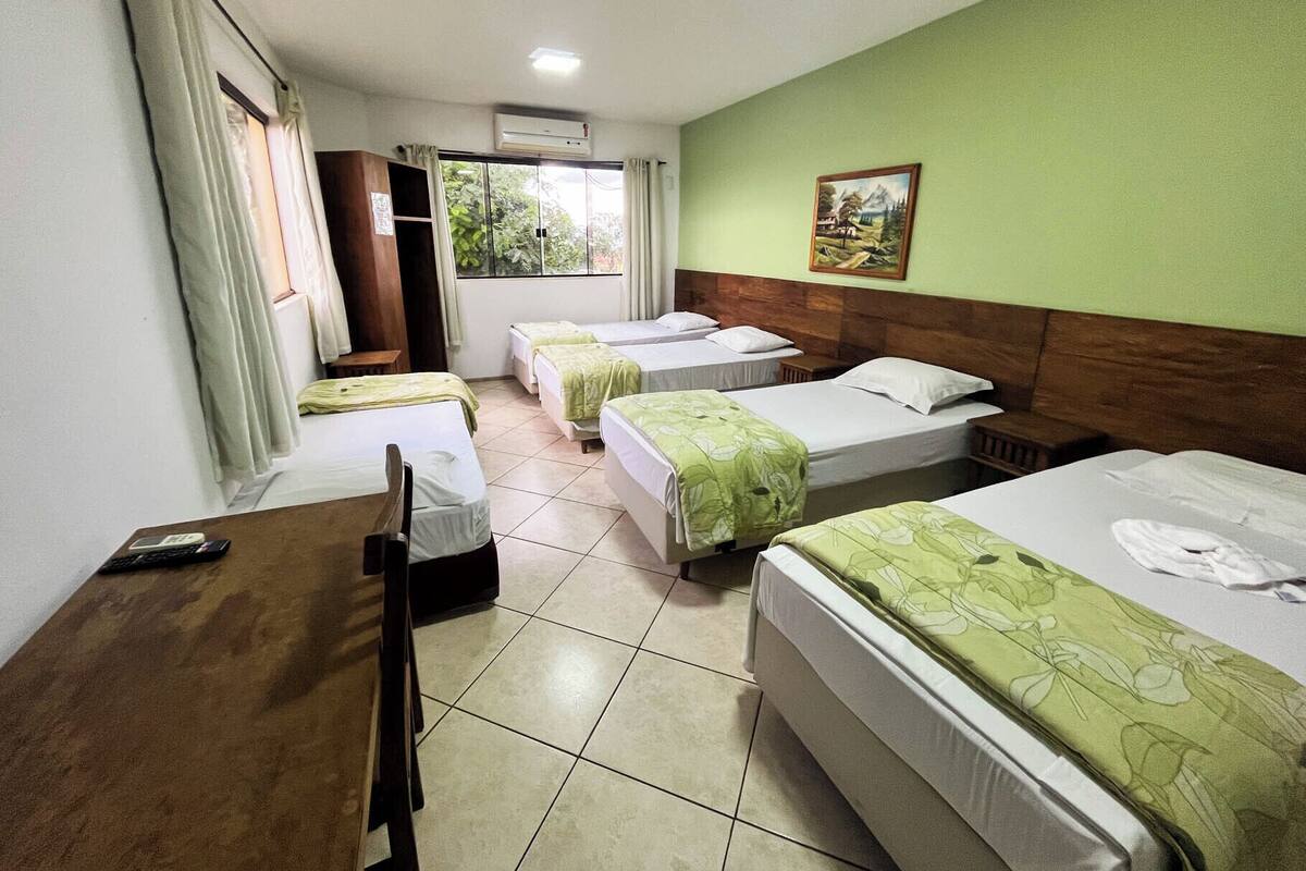 A hotel room with four single beds, a wooden desk, a chair, a window with curtains, and a painting on the wall.