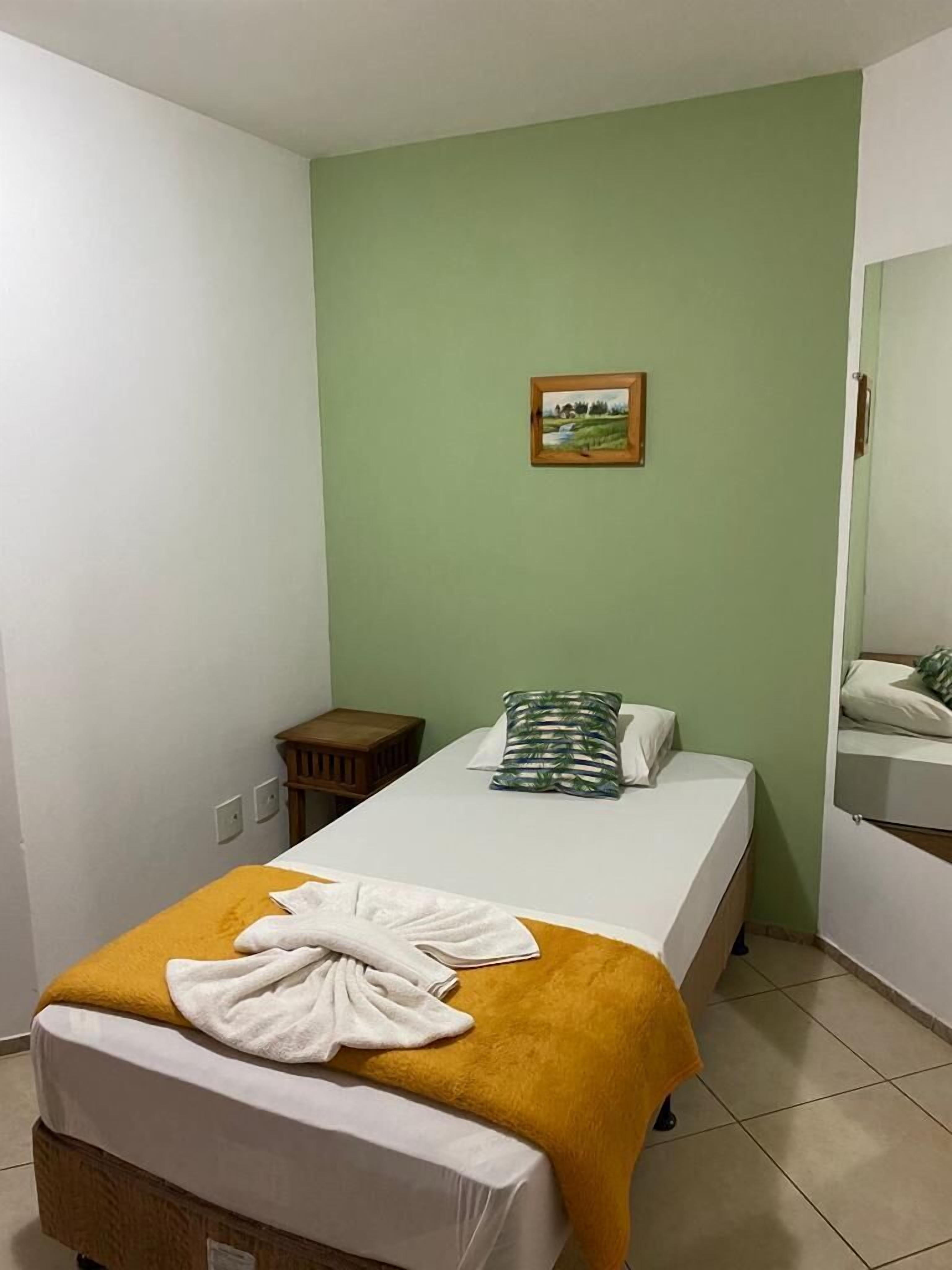 A single bed with a yellow blanket and a white towel, a small wooden nightstand, a green wall, and a framed picture above the bed.