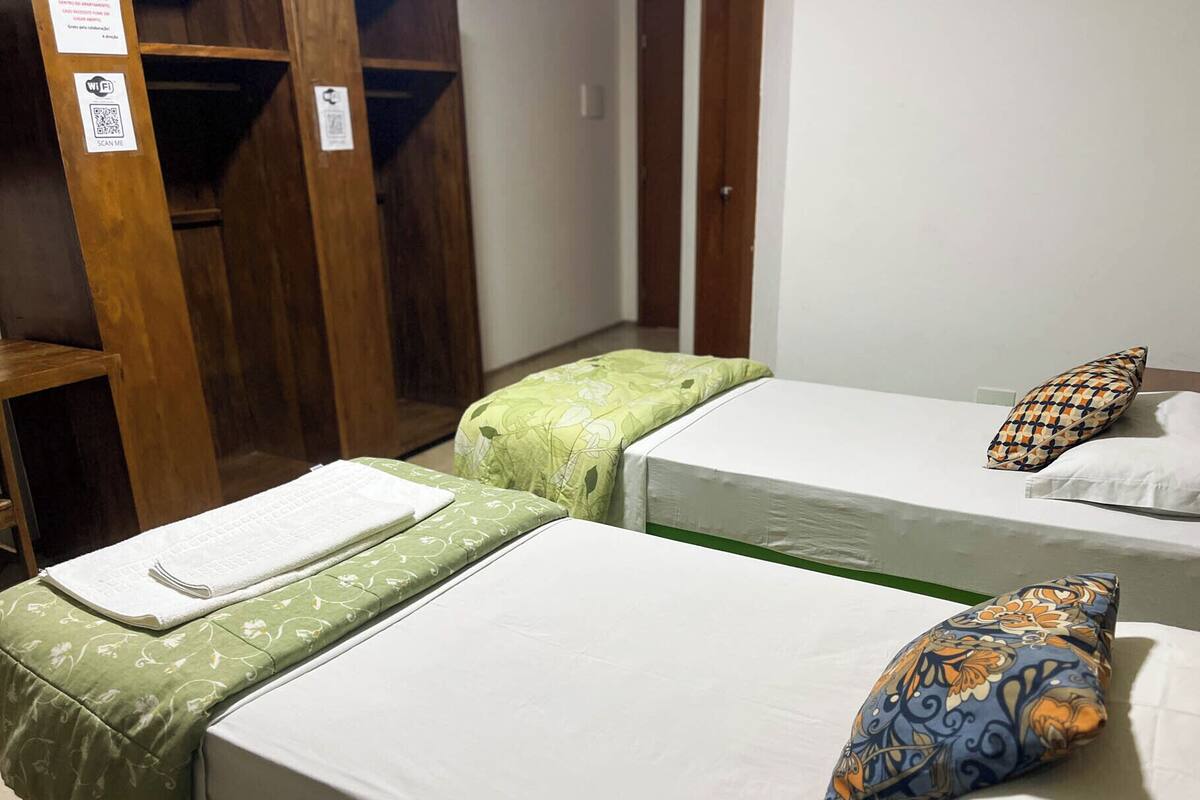 A room with two single beds, each with a pillow and a green floral bedspread. There is a wooden wardrobe and a door in the background.