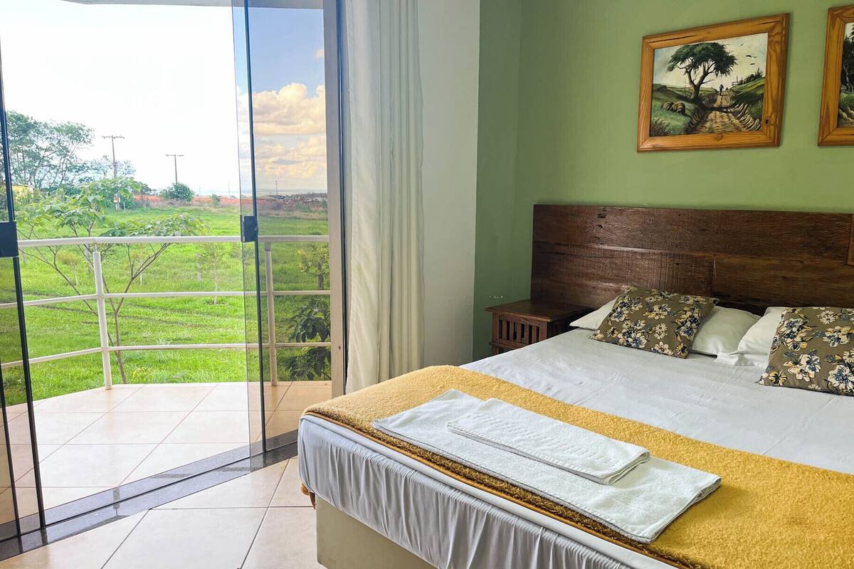 A bedroom with a large bed, a wooden headboard, and a view of a green landscape through a glass door.