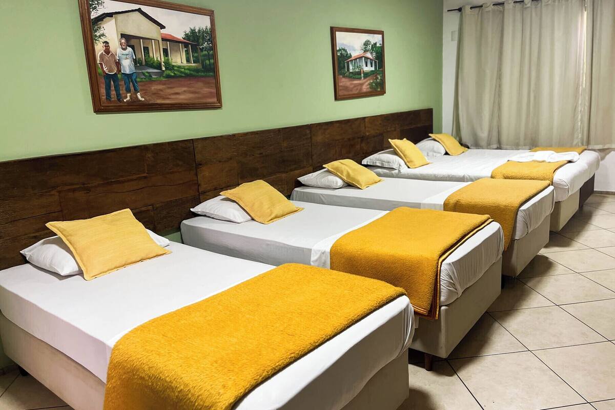 A hotel room with four single beds, each with a white mattress and a yellow blanket. The room has a wooden headboard, green walls, and two framed pictures on the wall.