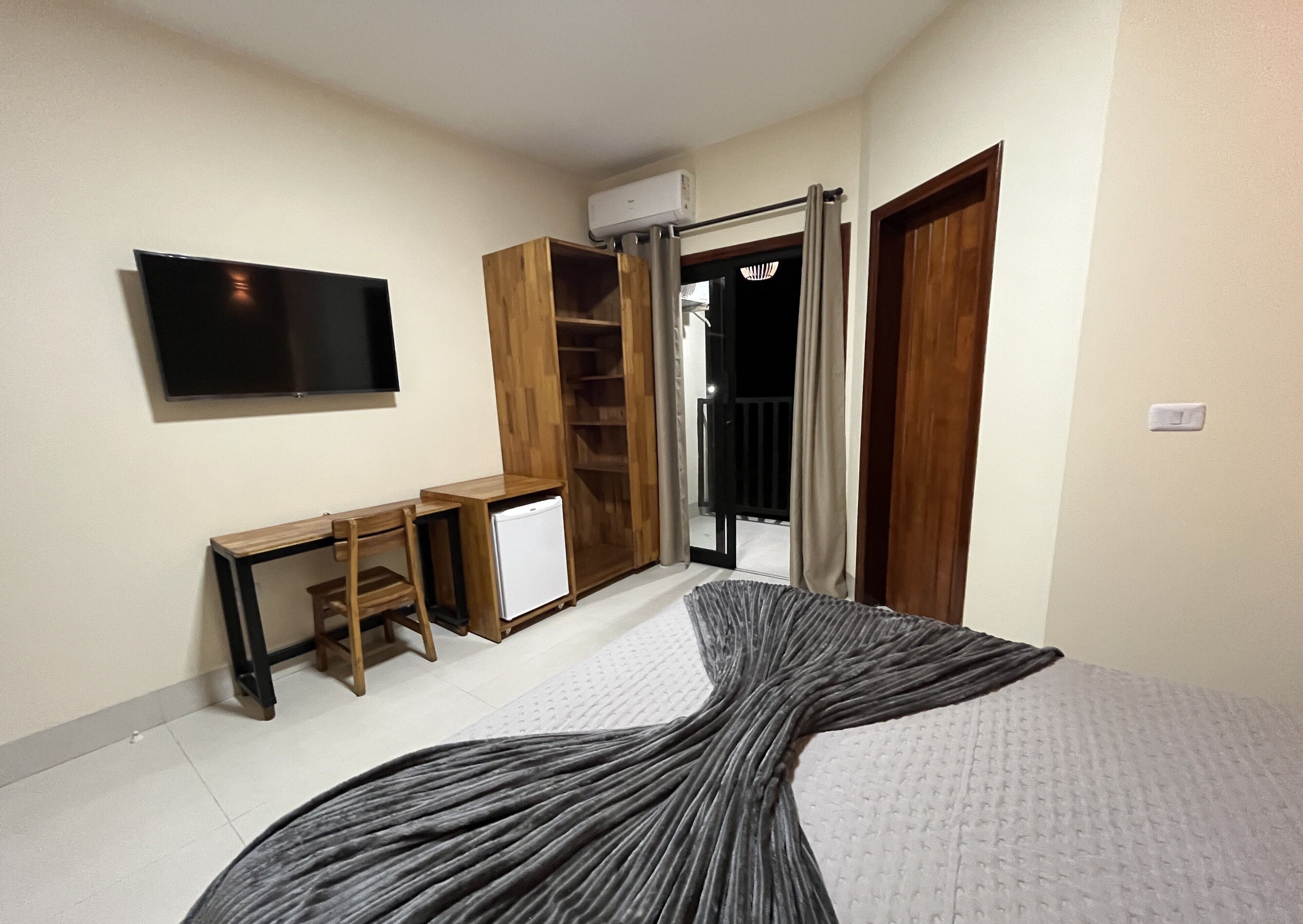 A bedroom with a bed, a TV, a desk with a chair, a wardrobe, and a balcony.