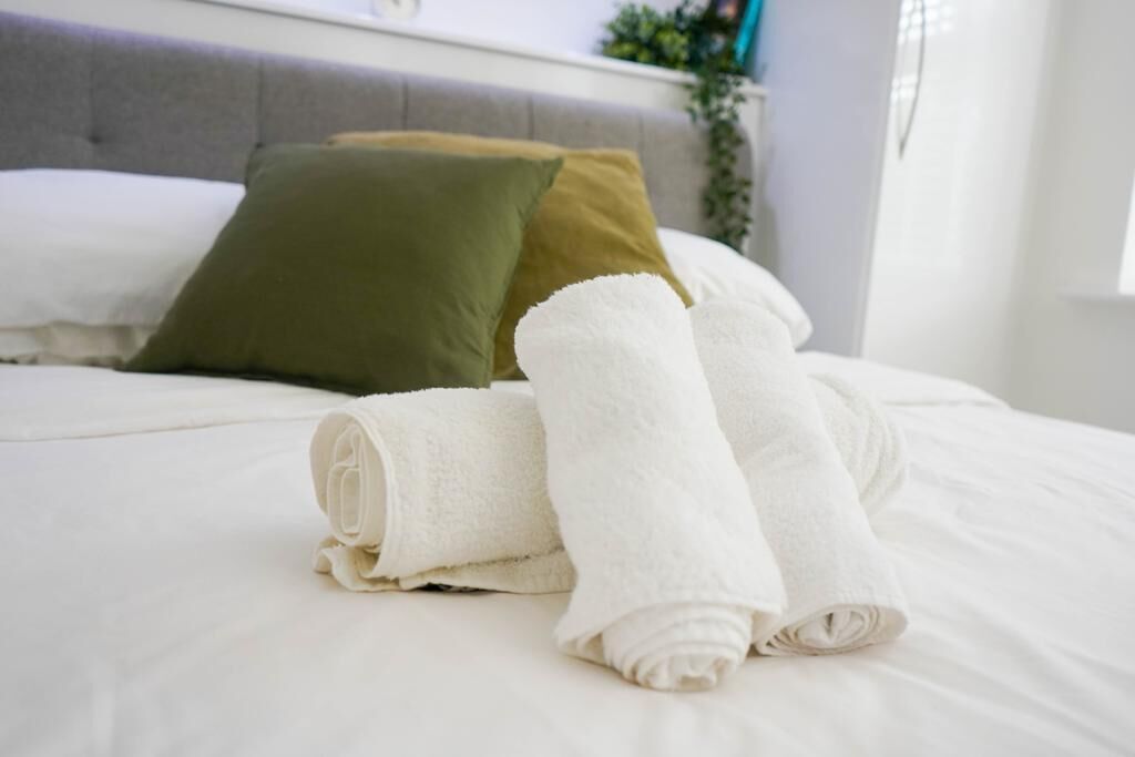 A neatly made bed with white linens and rolled towels, a grey headboard, and a green pillow.