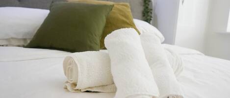 A neatly made bed with white linens and rolled towels, a grey headboard, and a green pillow.