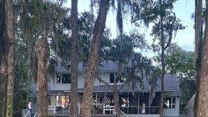 Une terrasse en bois menant à une maison dotée d’un porche, le tout entouré d’arbres et de verdure.