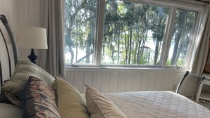 Une chambre à coucher avec un grand lit, une fenêtre donnant sur la verdure et une tête de lit agrémentée d’une lampe.