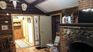 A rustic interior with a wooden floor, a fireplace, a television, and a door leading to a kitchen area. - Cozy Cabin (Clancy)