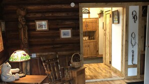 A rustic cabin interior with wooden walls, a rocking chair, a quilted bed, and a wooden table with a lamp. - Cozy Cabin (Clancy)