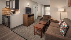 Habitación de hotel compacta con kitchenette, sofá con cojines estampados, una pequeña mesa de madera y televisión.