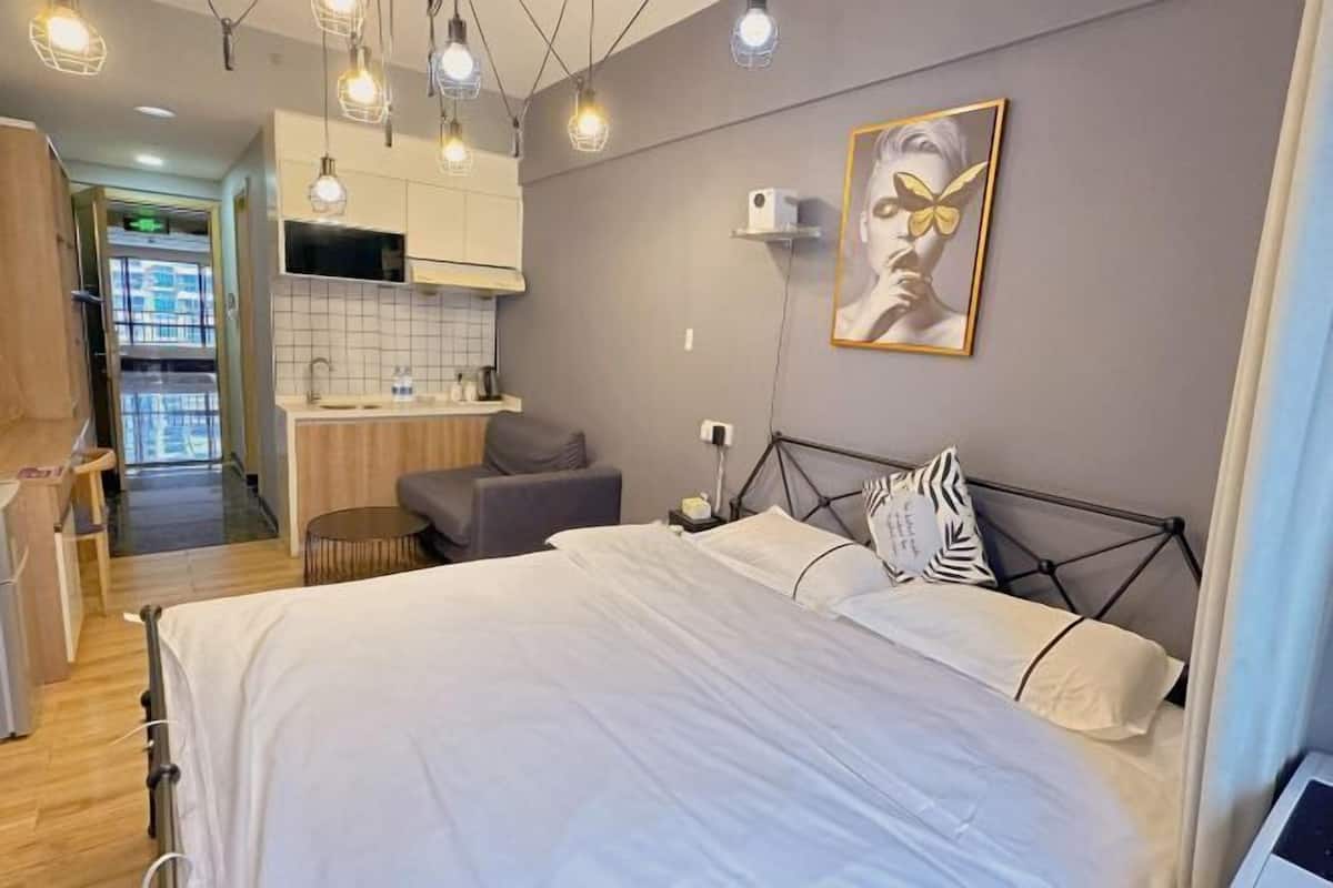 A modern hotel room with a bed, bedside table, and a kitchenette area.