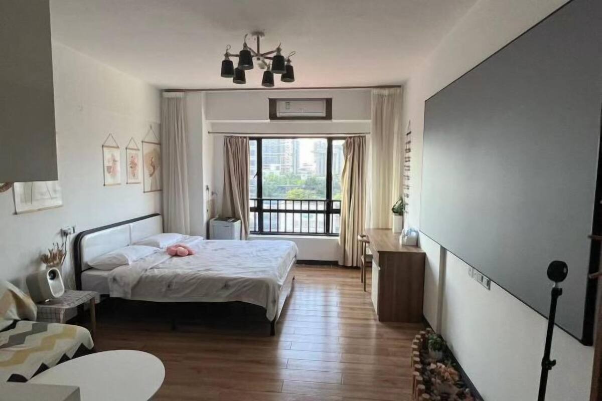 A bedroom with a bed, bedside table, chair, and a desk with a chair. There is a window with curtains and a view of buildings outside.