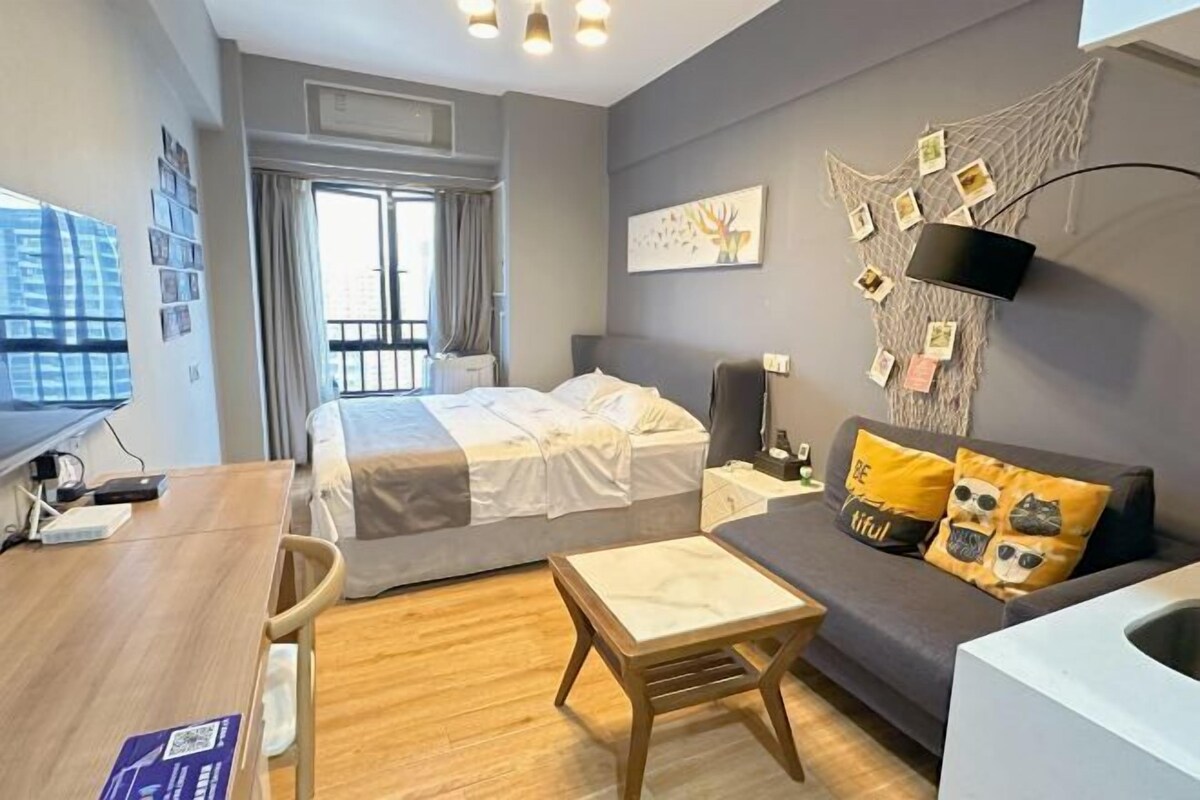 A modern hotel room with a bed, a sofa, a desk, and a small table. There is a window with curtains, a wall-mounted TV, and a wall with pictures and notes.