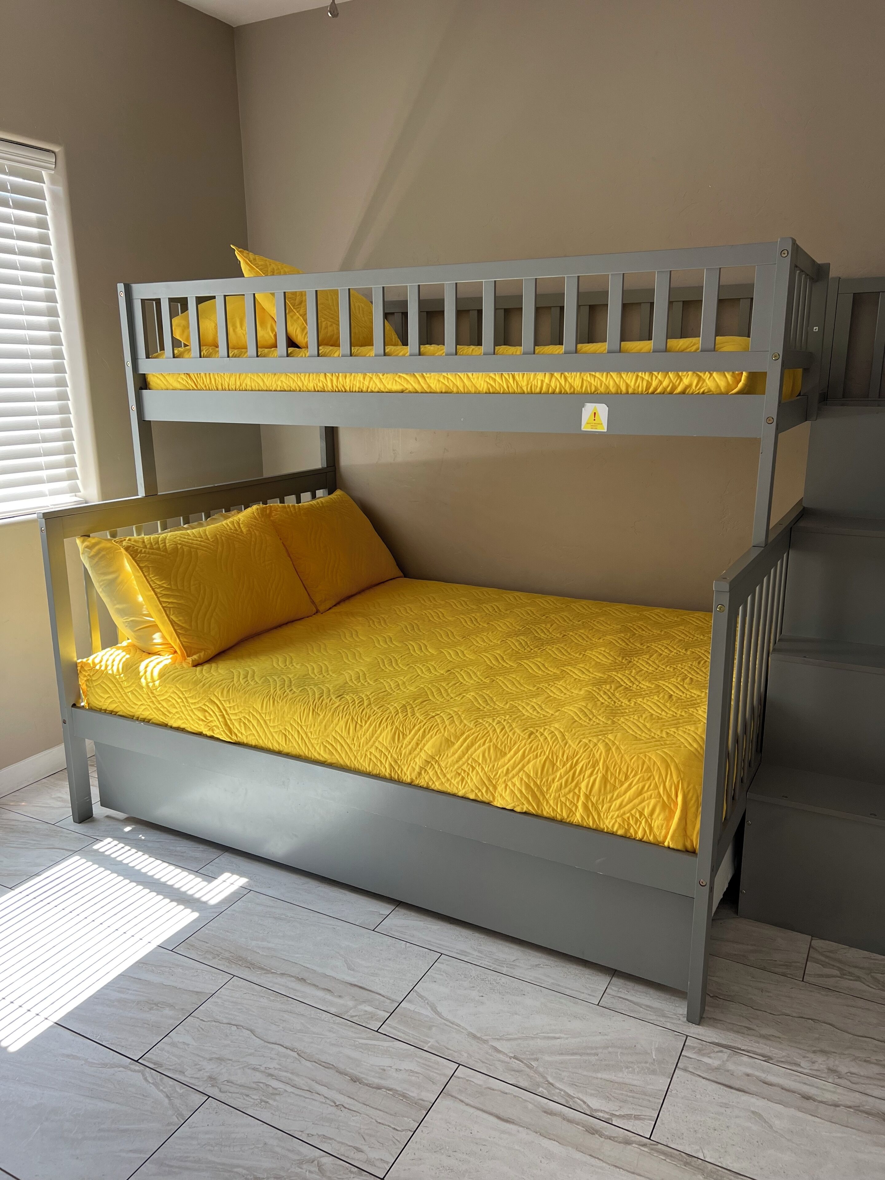 A bunk bed with a yellow mattress and pillows, grey metal frame, and a window with blinds.