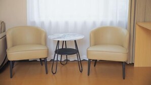 Een kamer met twee beige stoelen, een klein rond tafeltje en een raam met dunne gordijnen.