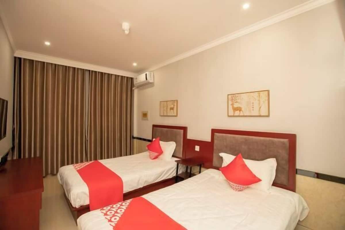 A hotel room with two single beds, each with a red accent pillow and a white bedspread. There is a wooden desk and a chair, and the room has a large window with curtains.