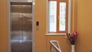 An elevator with a control panel, a window with a view of buildings, and a vase with flowers. - OYO Ulanhot Huide Business Hotel (Ulanhot)