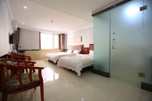 Quarto de hotel com duas camas, um guarda-roupa, uma escrivaninha com cadeiras e uma janela grande.