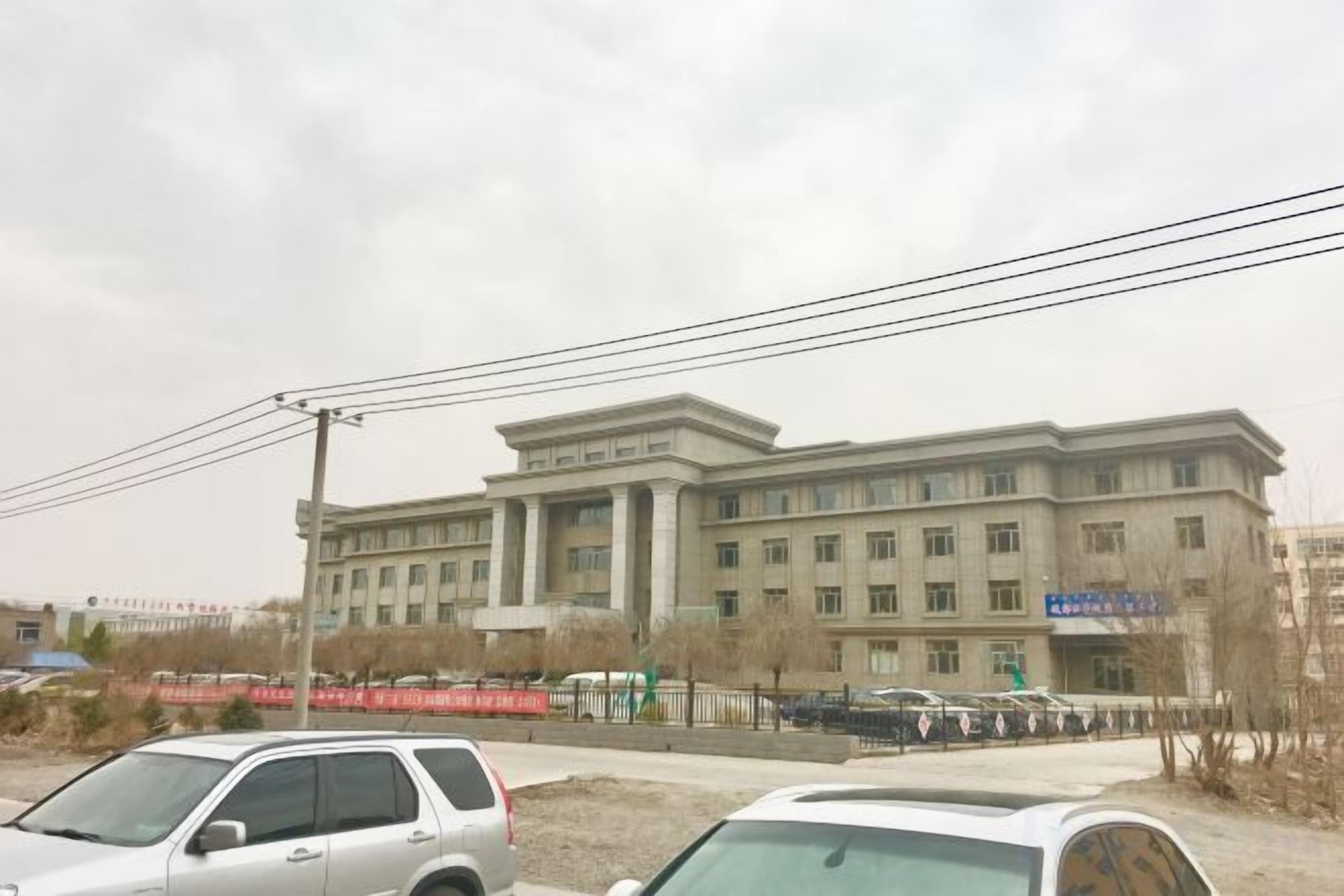 A large building with multiple windows and a flat roof, surrounded by parked cars and a fence.