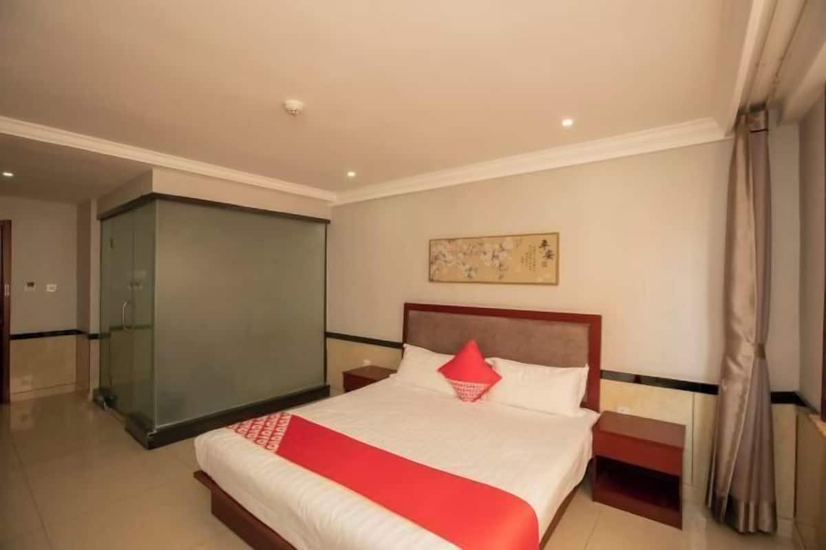 A hotel room with a large bed, a glass partition, a wooden nightstand, and a painting on the wall.